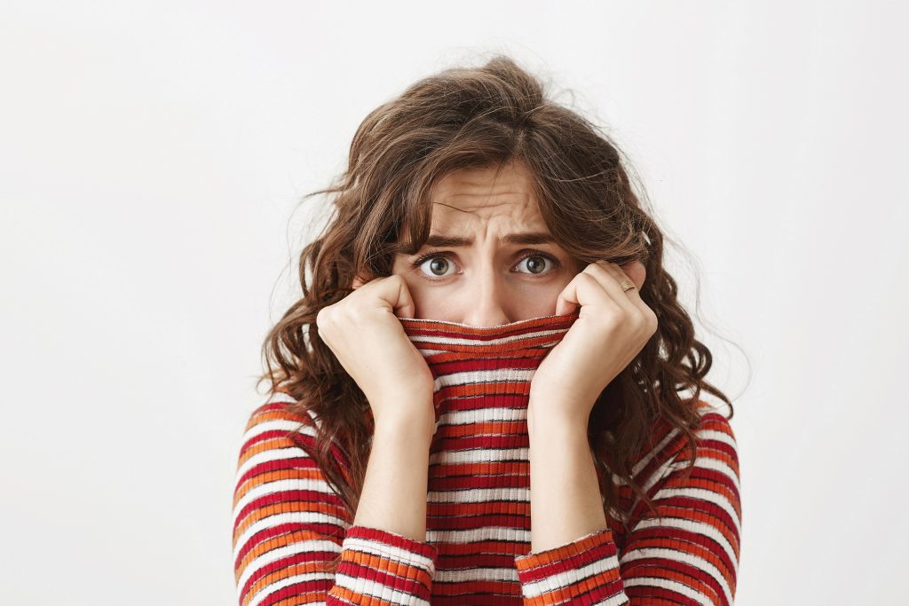 young female student hiding in sweater, looking through collar with afraid expression, frowning and wanting to cry while standing over gray background. 