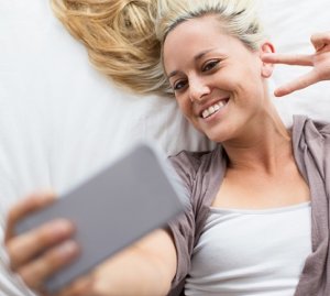 happy young woman lying in bed, taking selfie with smartphone