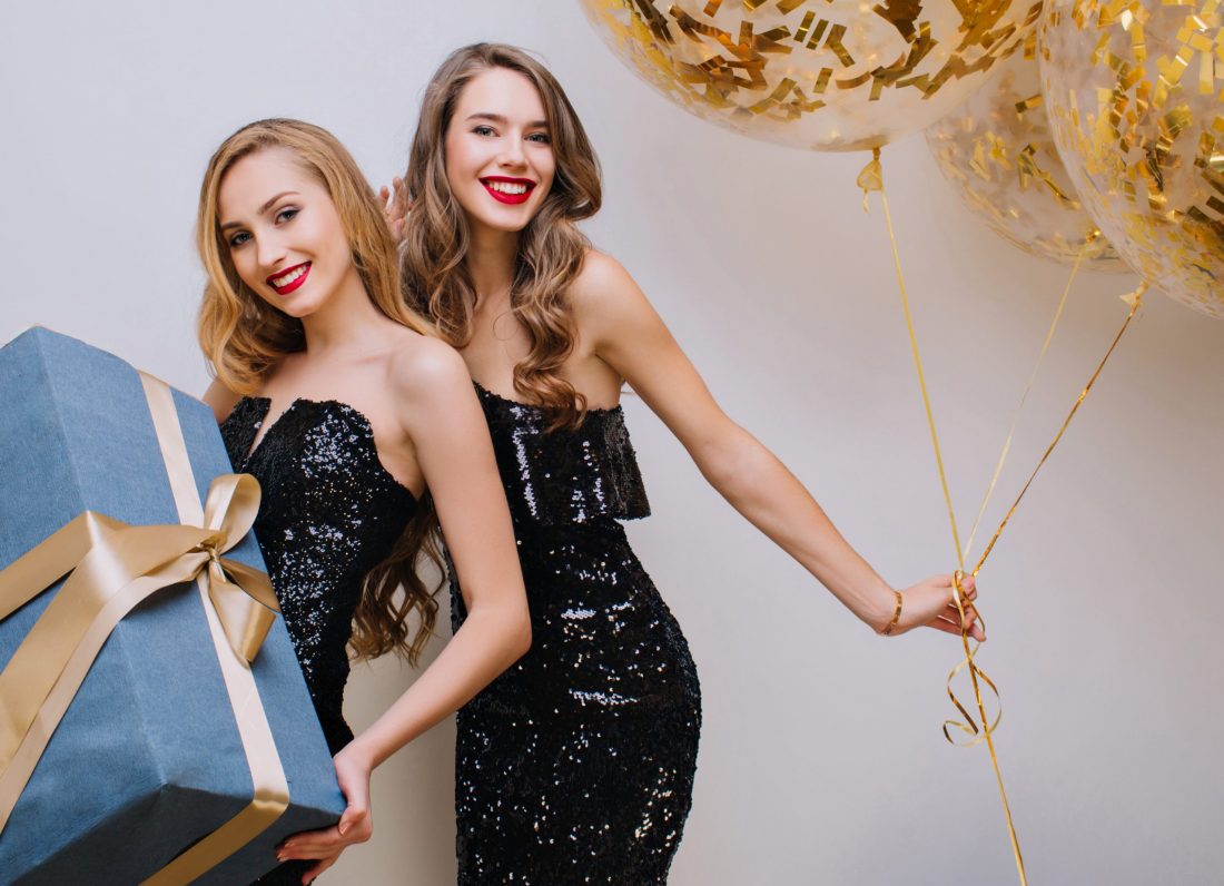 Fashionable curly european girl dancing with balloons behind blonde female friend. Indoor photo of joyful young woman holding present decorated with ribbon and gently smiling.. Fashionable curly european girl dancing with balloons behind blonde female friend. Indoor photo of joyful young woman holding present decorated with ribbon and gently smiling.